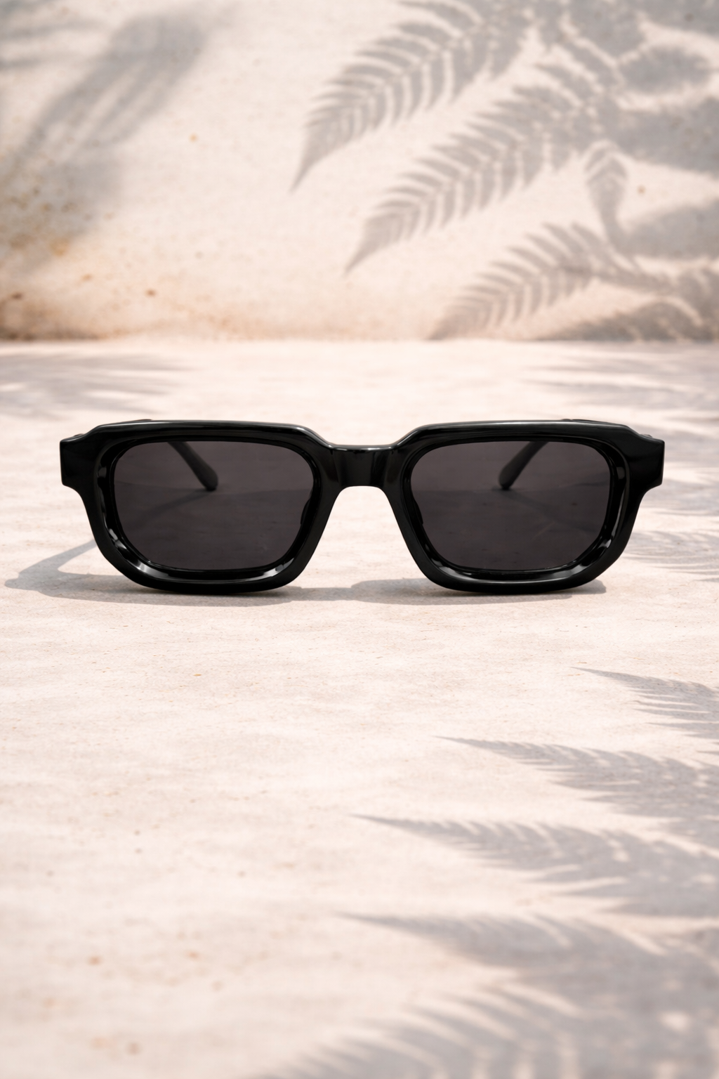 Black sunglasses on a sandy surface with palm leaf shadows
