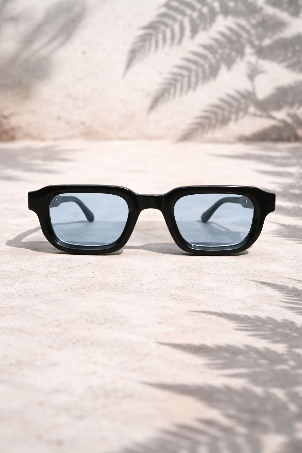 Black sunglasses with blue lenses on a sandy surface with palm leaf shadows